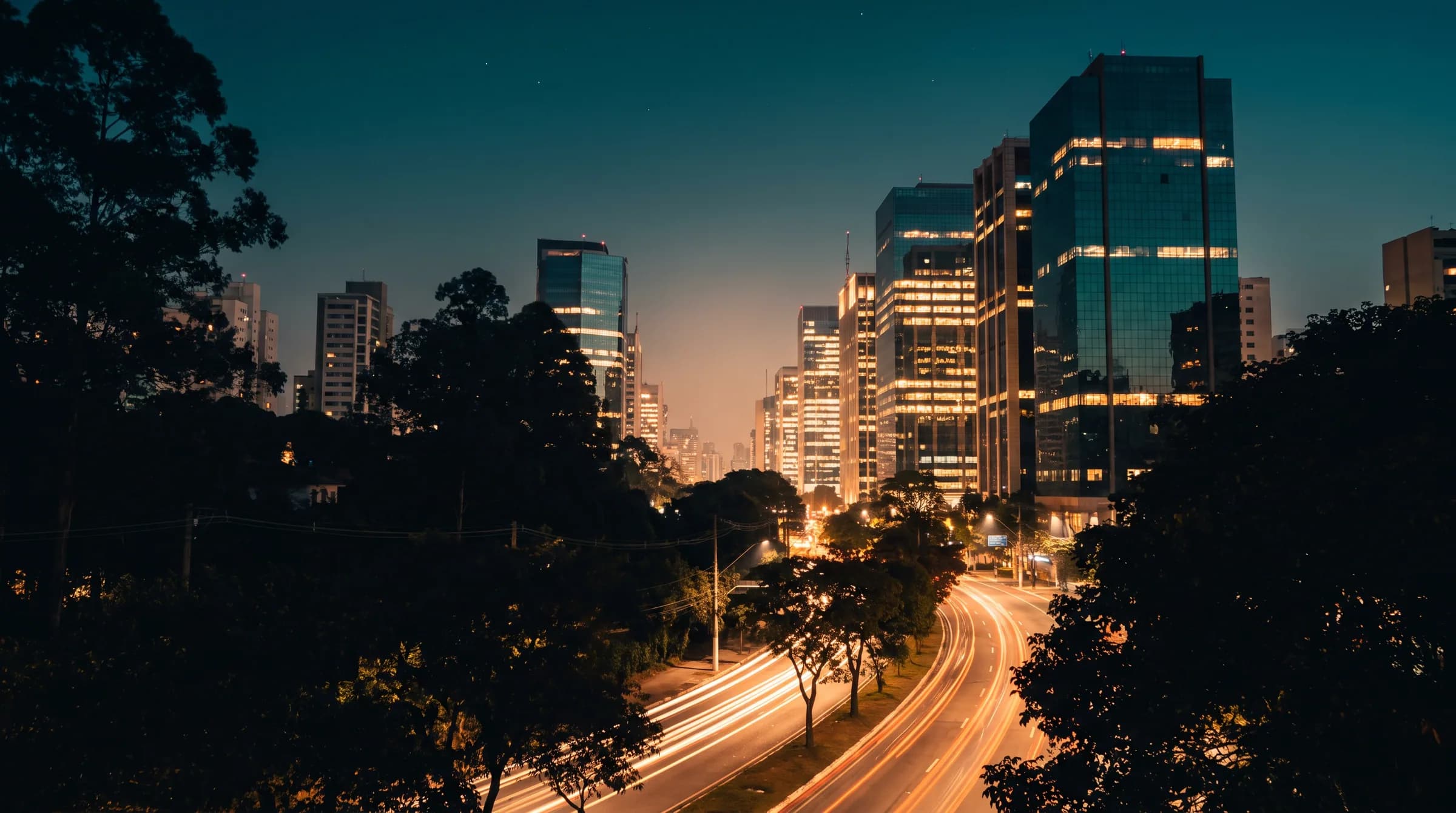 Avenida corporativa de São Paulo à noite, com torres de vidro iluminadas e rastros de luz do trânsito